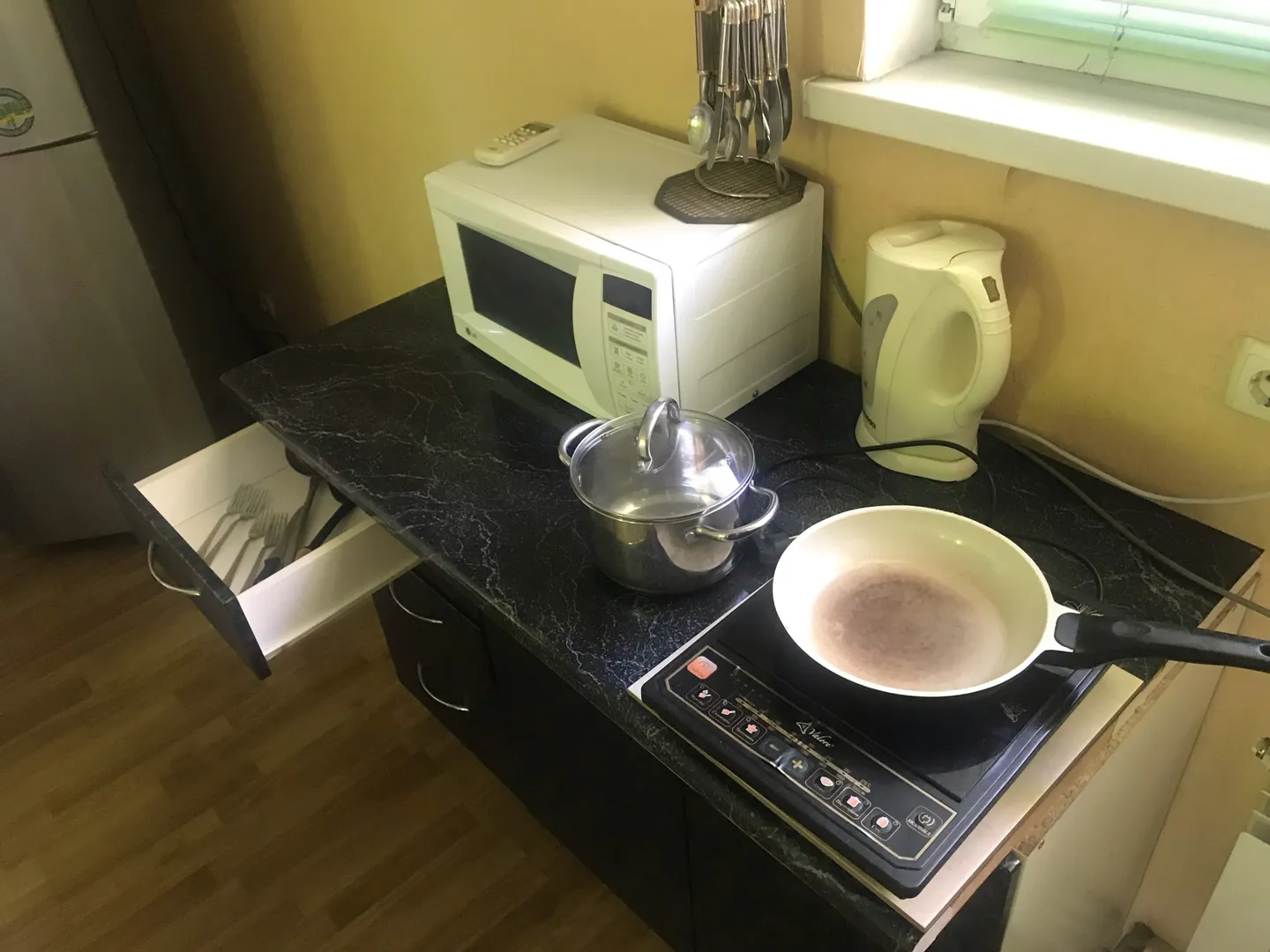 В кухонной зоне: микроволновая печь, индукционная лита с кастрюлей и сковородой, электрокипятильник, ложки, вилки, холодильник