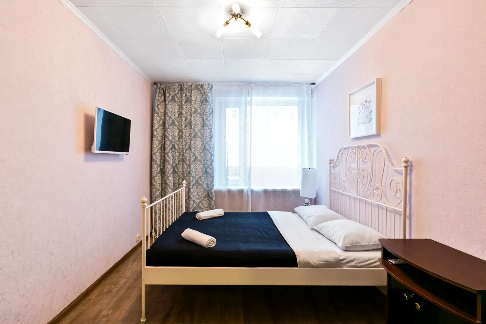 Спальня: крепкая железная кровать с чистым матрасом и застеленным бельем, ЖК-телевизор, плотные шторы и мягкий ковер.